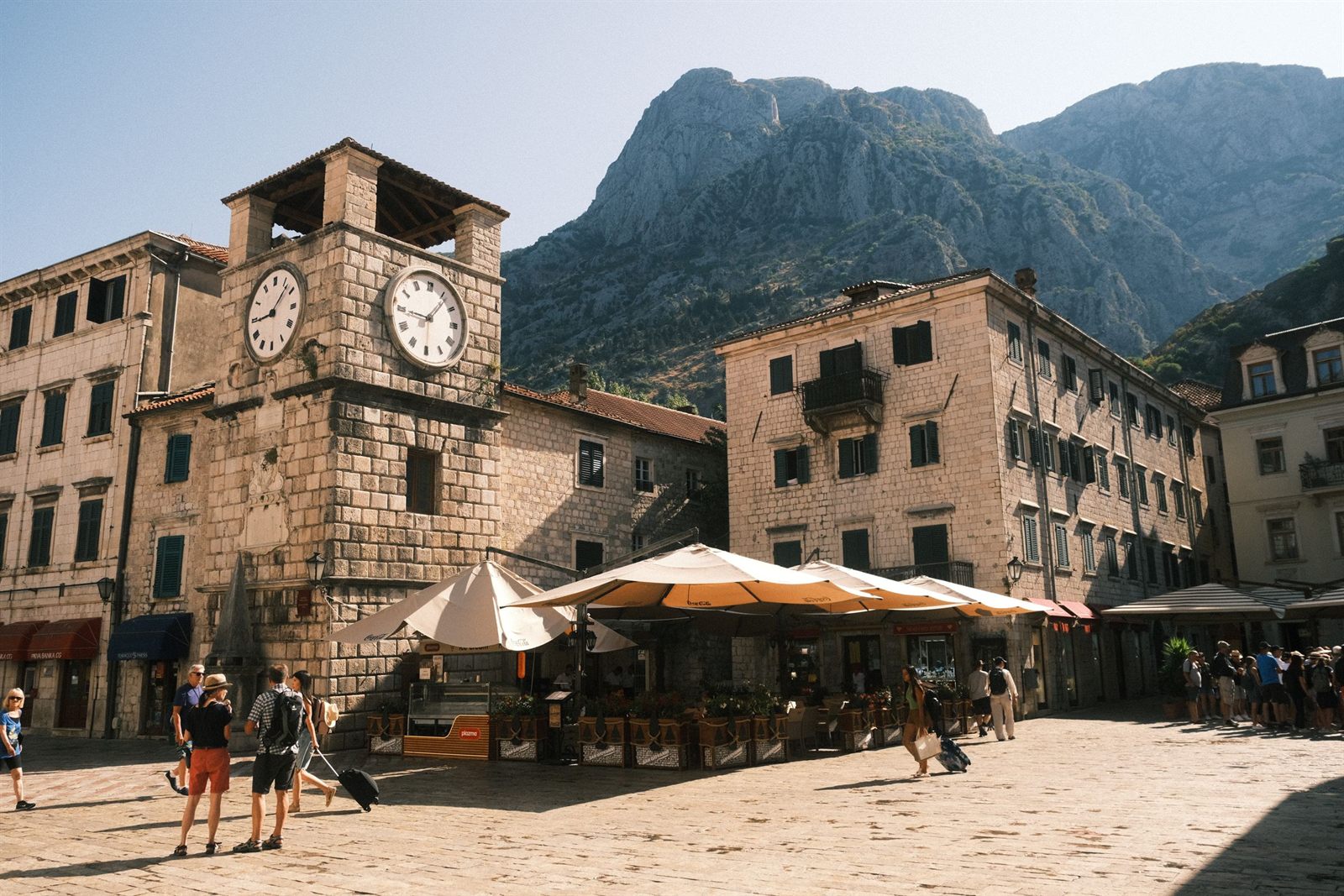 Clock tower and cafe tables in Kotor old town square below mountains